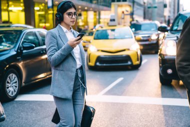 Gri takım elbiseli, gözlüklü, kulaklıklı, caddede yürürken cep telefonu kullanan modern bir kadın. 