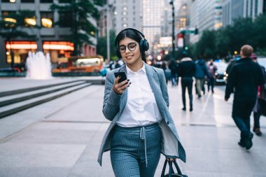 Şık giyinen, yuvarlak gözlüklü, akıllı telefon kullanan ve şehir merkezinde bulanık arka planda yürürken müzik dinleyen neşeli iş kadını.