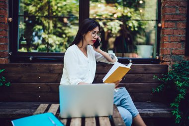 Klasik gözlüklü İspanyol serbest yazar, uluslararası cep telefonu konuşmaları sırasında proje detaylarını tartışmak için not defterinden bilgi kontrol ediyor, milenyum kadını hareketlilik bağlantısı kullanıyor