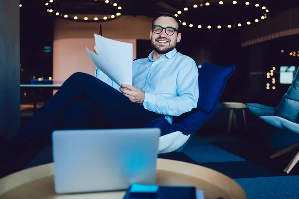 Young executive wearing glasses laughing and looking at camera with eyes half-closed while sitting in armchair in modern office and looking through documents