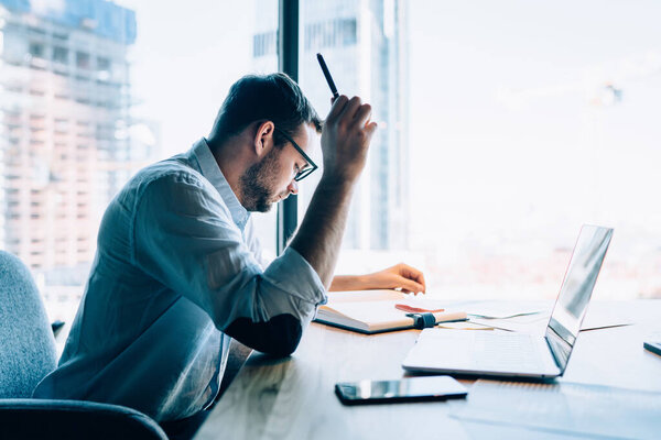 Concentrated smart intellectual male in glasses with pen in hand in blue shirt checking notepad  in office sitting at table with laptop opposite of window