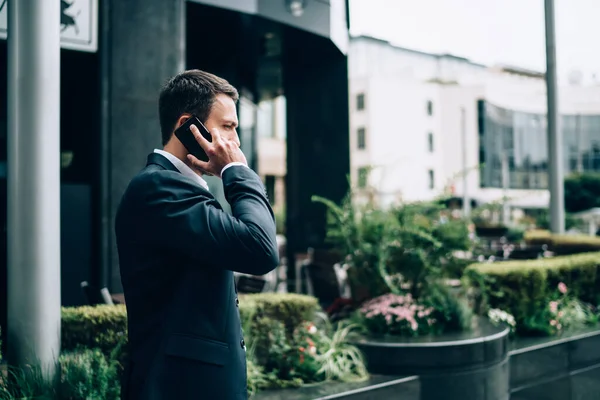 站在花坛边接电话 在现代城市街道上用智能手机交谈的成年男性经理侧视图 — 图库照片