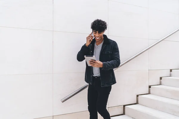 現代的なスタイリッシュな明るい笑顔若いブルネットアフリカ系アメリカ人男性カジュアルな服装で電話で話し 昼間の街の階段に立っている間にタブレットを使用して — ストック写真