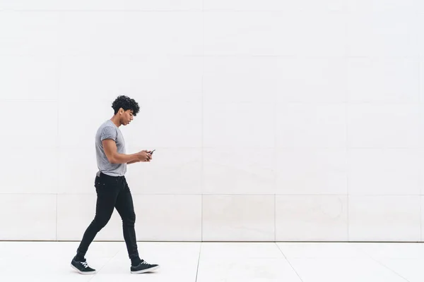 Modern adult fit male curly ethnic in gray shirt and black jeans surfing mobile while walking alone on street in front of white marble wall