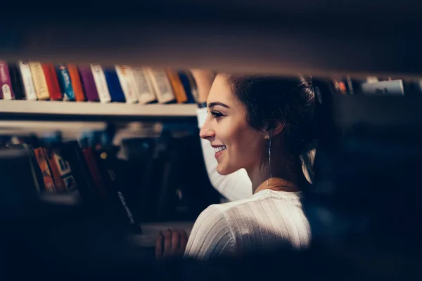 从书架侧面看穿着白色夹克的少女在选择图书馆看书时的笑容 — 图库照片