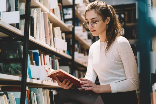 スタイリッシュな眼鏡と本棚の近くに座って図書館で本を勉強するカジュアルな服で快適な黒い髪の女性のティーンエイジャー — ストック写真