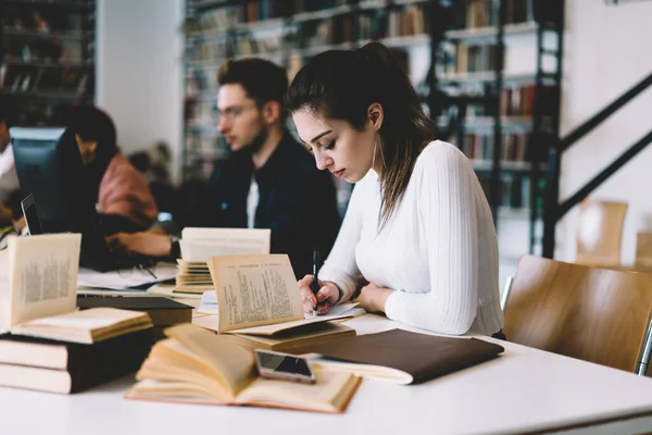 专心致志的年轻女性 穿着休闲装 在大学的一个项目上工作 她坐在图书馆的书桌边看书 写笔记 — 图库照片