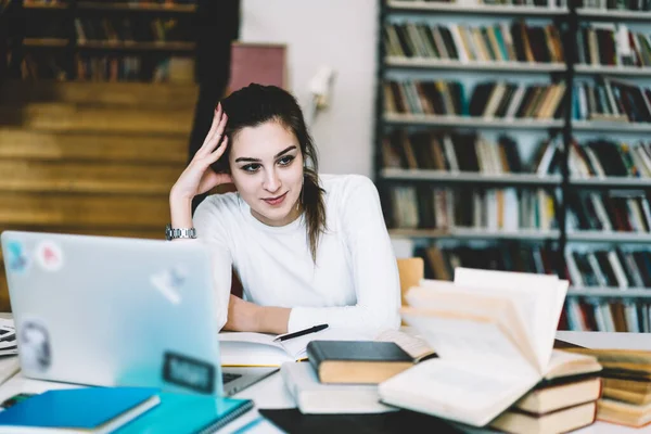 心满意足的黑头发女学生穿着白色夹克坐在桌旁 靠在手上做作业 带着笔记本电脑和书本在图书馆里看书 — 图库照片