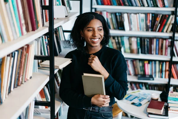 African American joyful smiling lady in casual clothes buying book for homely reading enjoying spending time standing about bookshelf in library