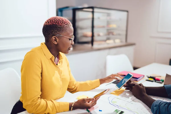 Side view of modern African American female entrepreneur sitting with mobile phone in hand and looking at smartphone of crop colleague during business meeting in cafeteria