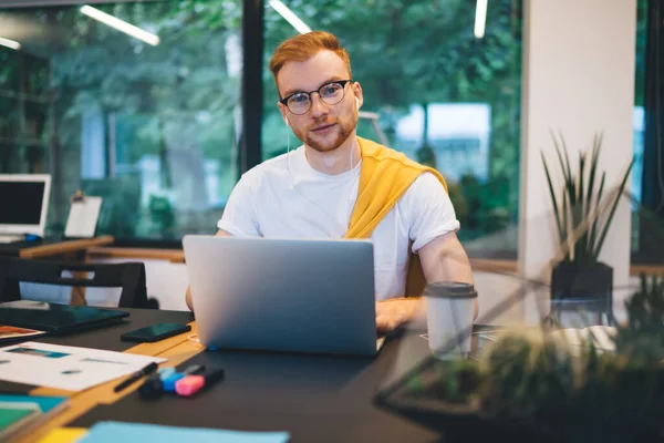 年轻而严肃的红头发男性 身穿休闲装 穿着白色T恤 坐在笔记本电脑旁 边喝咖啡边浏览 — 图库照片