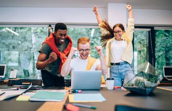 Group of excited diverse young people clenching fists and celebrating successful project fulfillment while working in modern creative workspace together