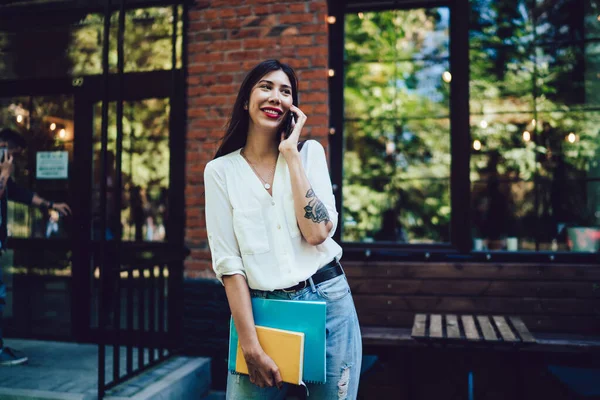 Cheerful Latino female university student with sketchbook standing at urbanity and using cellphone gadget for making roaming mobile call and discuss project details, international connection concept