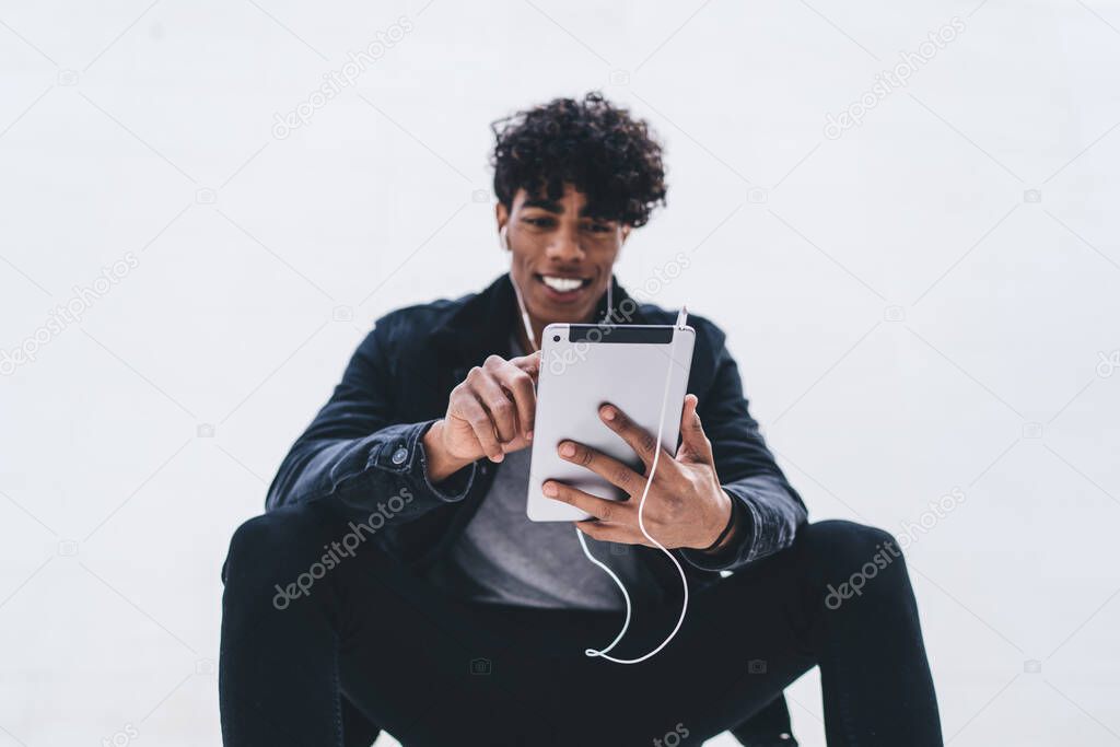 From below of cheerful African American male wearing black clothes choosing app on tablet listening to music on white background