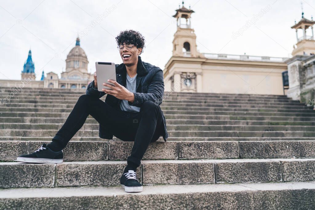 Laughing curly modern male in black jacket using tablet for video chat while sitting on street stairs on blurred background