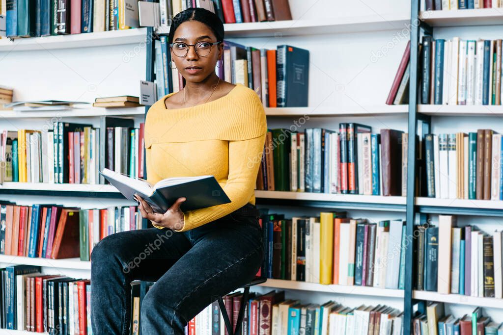 Serious African American lady in casual outfit and glasses looking away while sitting on ladder and reading book in library