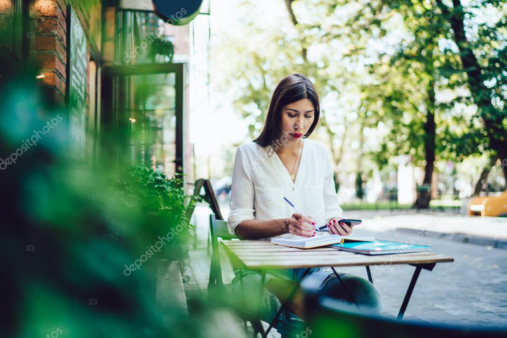 Attractive female blogger reading received email on cellular connected to 4g wireless for browsing internet during learning in street cafeteria, hipster girl chatting researching information for test