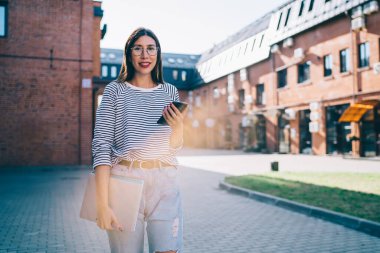 Teknoloji donanımlı neşeli kız öğrencilerin yarım boy portresi. Açık havada durup kameraya gülümsüyor. Cep telefonuyla 4G 'ye bağlanan mutlu hippi kız.