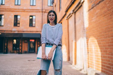 Serbest çalışmak için modern dizüstü bilgisayarı tutan ve boş zamanlarında açık havada kameraya bakan gündelik kıyafetli başarılı bir kadının yarım boy portresi.