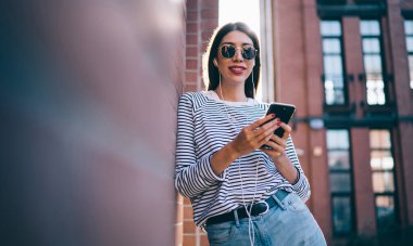 Modern cep telefonuyla radyo uygulaması, pozitif kadın kulaklıkları test eden modern cep telefonuyla şehir merkezinde arkadaşının müzik podcasının keyfini çıkaran bin yıllık hipster kızının yarım boy portresi.