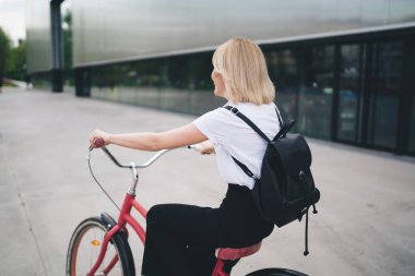 Gündelik kıyafetli, siyah sırt çantalı, şehir caddesindeki binalara karşı bisiklete binen ve başka yöne bakan modern kadınların arka plan görüntüsü. 