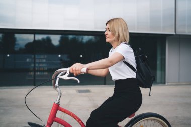 Şık, modern sarışın bir kadının, kısa saçlı, pembe eski bir bisiklete binip şehir merkezindeki binadan uzağa bakmasının yan görüntüsü. 