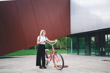 Gülümseyen modern sarışın kadın, sıradan kıyafetler içinde, siyah sırt çantalı, şehir merkezindeki pembe bisikletli, başka yöne bakan. 