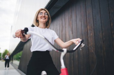 Gülümseyen milenyum kadınının düşük açılı görüntüsü. Tişört ve siyah pantolon, ileriye bakarken koyu kereste duvarda kırmızı bisiklet sürüyor.