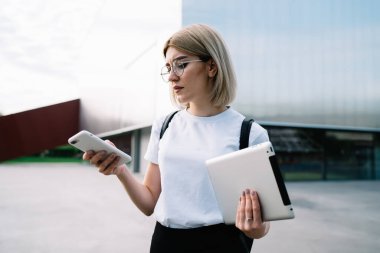 Gözlüklü, günlük giysiler içinde dikkatli bir kadın. Ofis binasının yanındaki caddede dikilirken konsantrasyonuyla akıllı telefona göz atıyor.