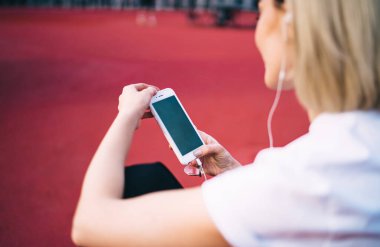 Yüzsüz, modern, gündelik giysiler içinde, lastik spor sahasında otururken arkadaşlarıyla mesajlaşmak için cep telefonu kullanan bir kadın.