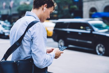 Elektronik kulaklıklarla iş rehberini dinlerken cep telefonuyla bilgi kontrol eden beyaz erkek tüccar, uygulama üzerinden ortağa göndermek için e-posta cevabı yazan deneyimli bir adam