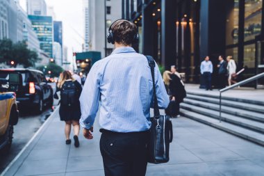 Deri çantalı, resmi giyinmiş, finans bölgesindeki iş toplantılarına elektronik kulaklıklardan ses kitabı dinleyerek giden zeki bir erkek yöneticinin arka planda görüntüsü.