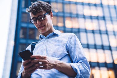 New York 'ta ışıklandırılmış yüksek binaya karşı telefonda mesajlaşan tişörtlü ve gözlüklü çağdaş adamın alçak açısı.