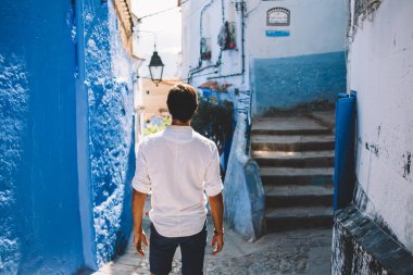 Fas 'ın yaşlı bir kasabasında güneşli bir günde dar sokaktaki eski evlerin önünde duran beyaz gömlekli, isimsiz bir adamın arka görüntüsü.