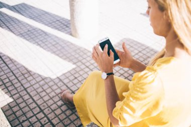 Yukarıdan bakıldığında, beyaz perdeye odaklanan sarı elbiseli sakin sarışın kadın ve şehirdeki güneş ışınları arasında gri kaldırım ve sütunlara karşı akıllı telefonla etkileşim içinde.