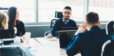 Konferans odasında dizüstü bilgisayarla otururken şirket projesini tartışmak için çalışanlarla işbirliği yapan neşeli, kendinden emin, zeki bir uzman.