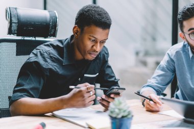 Modern ofisteki önemli bir iş toplantısında kalem taraması yapan akıllı telefonlu ve düşünen Afro-Amerikalı bir adam.