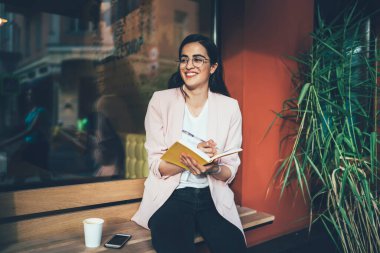 Gözlüklü, şık elbiseli, içecek ve akıllı telefonlu mutlu bir bayan kahverengi ahşap bankta oturmuş, notlarda modern kafe ve şehirdeki yeşil bitkilerin bulanık cephelerine karşı yazarken ve gülümserken.