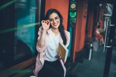 Yüksek açılı, zarif elbiseli, not defteri takan, gözlüklü ve şehirde ahşap bankta dinlenirken kameraya gülümseyen hoş bir yönetici kadın.