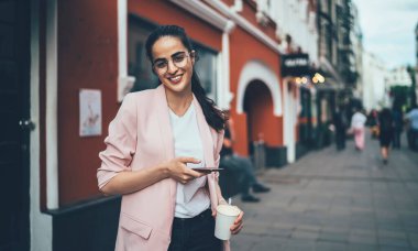 Gözlüklü, beyaz tişörtlü, pembe ceketli, sokakta dikilirken kameraya gülümseyen ve şehir merkezindeki eski renkli binaların bulanık dış yüzlerine karşı akıllı telefon kullanan kendine güvenen koyu renk saçlı kadın.