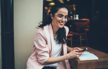 Keyifli, genç ve çekici bir bayan günlük kıyafetler ve gözlüklerle cep telefonu kullanıyor kafeteryada dinleniyor ve kameralara bakıyor.