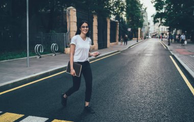 Beyaz gömlekli, siyah pantolonlu, spor ayakkabılı, yaya geçidinde yürürken kameraya gülümseyen ve şehir görüşünü bulanıklaştıran akıllı telefon kullanan bin yıllık mutlu öğrenci.