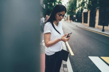 Sakin bir milenyum çalışanı gözlüklü ve günlük kıyafetleriyle ekranda yoğunlaşan kağıtlarla ve akıllı telefonuyla yaz günü kaldırım kenarında bulanık caddeye karşı dikilirken iletişim kuran.