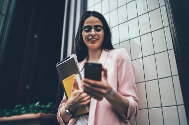 Elinde kağıtlar ve günlükle bina duvarının yanında duran ve internette cep telefonundan haber okuyan başarılı, modern meşgul bir kadından. 