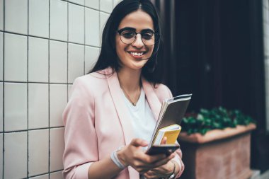 Neşeli, kendine güvenen, gözlüklü ve spor ceketli bir kadın. Elinde kağıt klasör, organizatör ve cep telefonu ile şehrin modern binasının duvarının yanında duruyor.