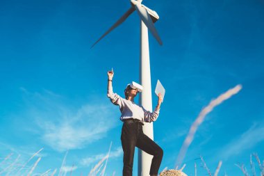 VR gözlük takan genç bir bayan imarcı güneşli bir günde yel değirmeninin yanında dikilirken dokunmatik program ve stil kullanarak sanal siber uzay dizayn ediyor.