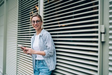 Sıradan kıyafetler giyen, moda gözlüklü, gülümseyen, kameraya bakan, pencereye yaslanan ve akıllı telefon kullanan neşeli genç bir kadın.
