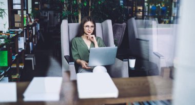 Çekici, serbest çalışan genç bir kadın dizüstü bilgisayarını dizlerinin üzerine çöküp rahat koltukta oturmuş elinde çenesiyle kütüphane odasına yaslanmış özenle kameraya bakıyor.