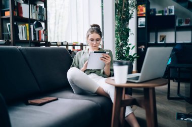 Serbest giyinen genç ve güzel bir bayan kütüphanede koltukta oturup tablet kullanarak internetten bilgi arıyor.