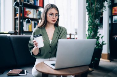 Özenli düşünceli bir kadın tablet ve not defteriyle rahat bir koltukta oturuyor ve arka planda bulanık bir şekilde dizüstü bilgisayar kullanarak kahve içiyor.